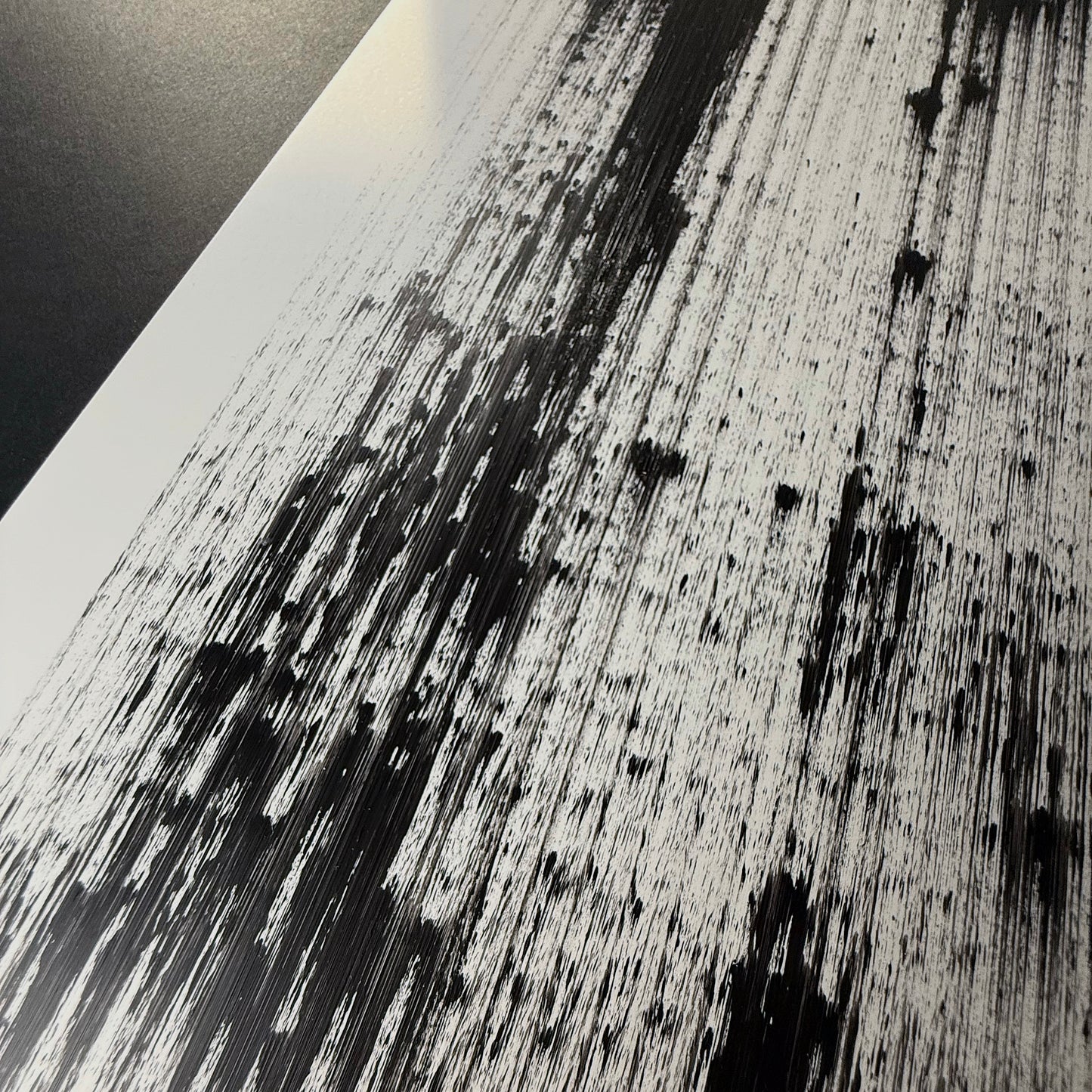 Close-up shot of a black abstract painting, acrylic on white Yupo synthetic paper, made with a pen plotter dragging black acrylic paint in repeated and constant vertical gestural strokes. Shot at a steep raking angle, the surface reads as a fine spray of black spatter and hairline streaks scattered over the white, denser toward the lower left and fading to airy specks at the top.