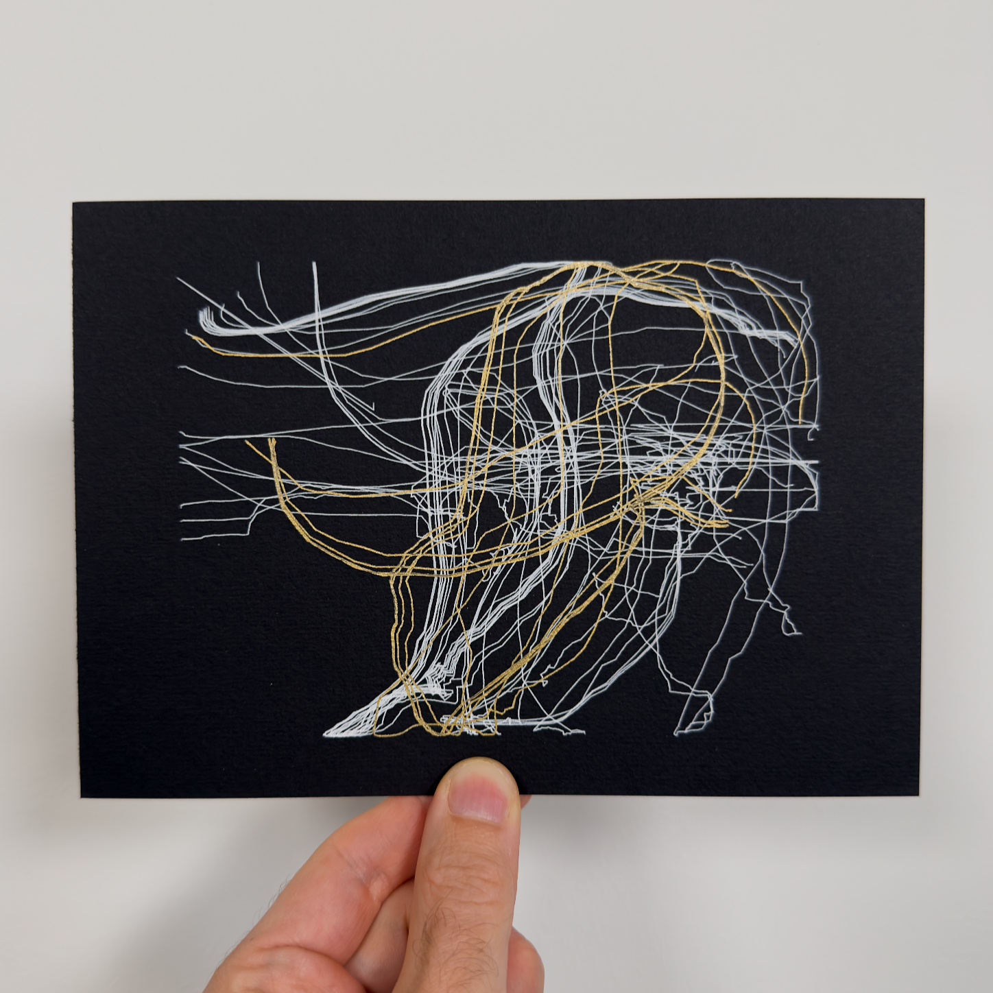 Hand holding a black card with white and gold string art design on a light gray background